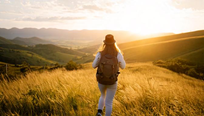 A woman wearing a hat and carrying a backpack walks through a golden field at sunset, with rolling hills and a wide valley stretching into the distance