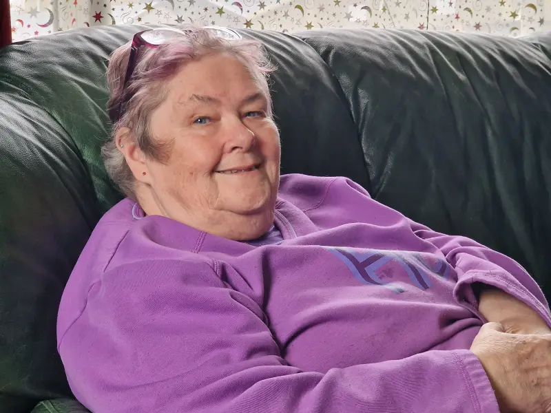 Mum smiling gently while relaxing on a dark green sofa, wearing a purple sweatshirt and glasses on her head