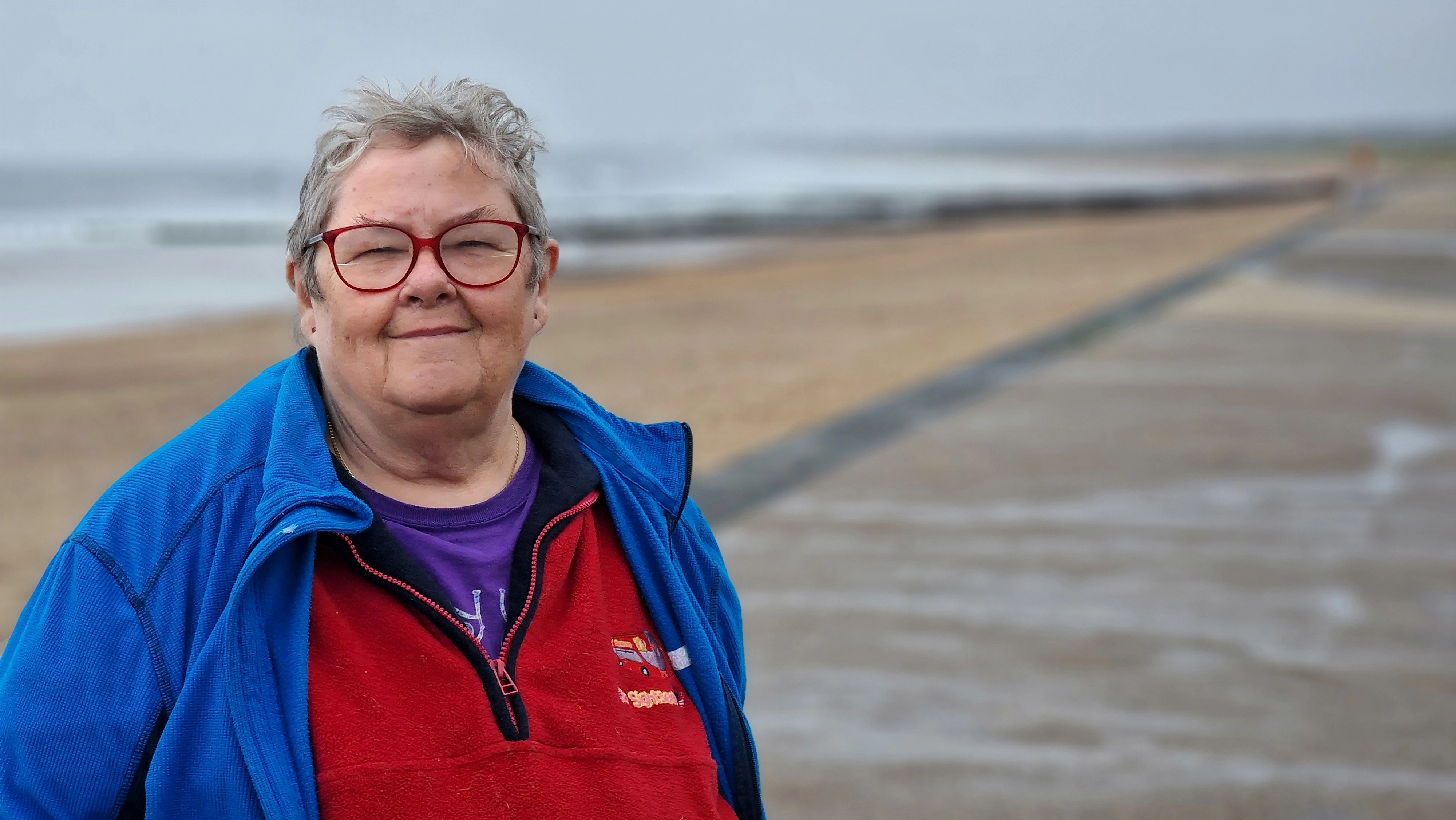 Mum standing on a misty beach, wearing red glasses and a colourful fleece, looking calm and content.
