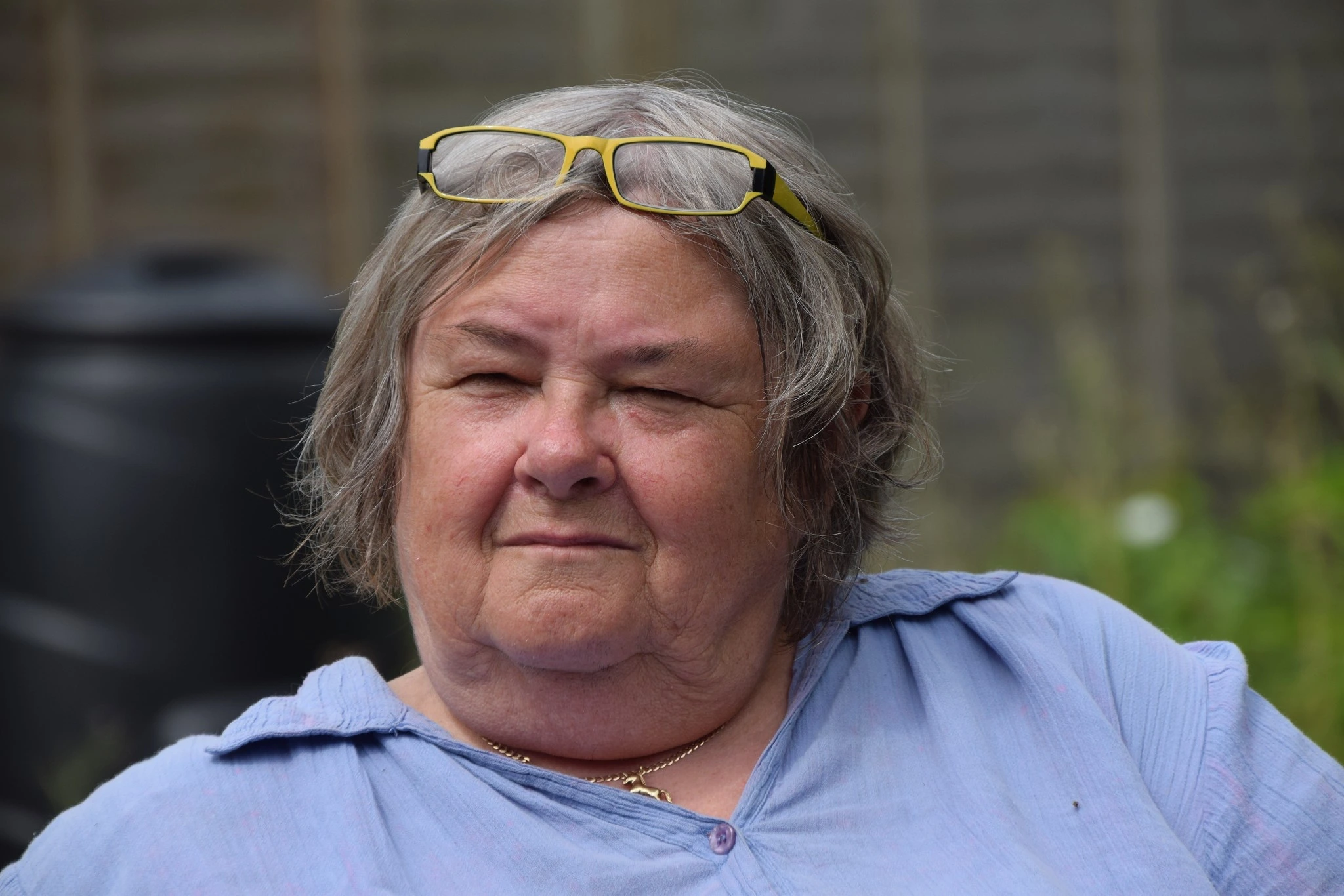 Close-up of Mum outdoors, squinting slightly with a thoughtful expression, wearing light blue and glasses on her head.
