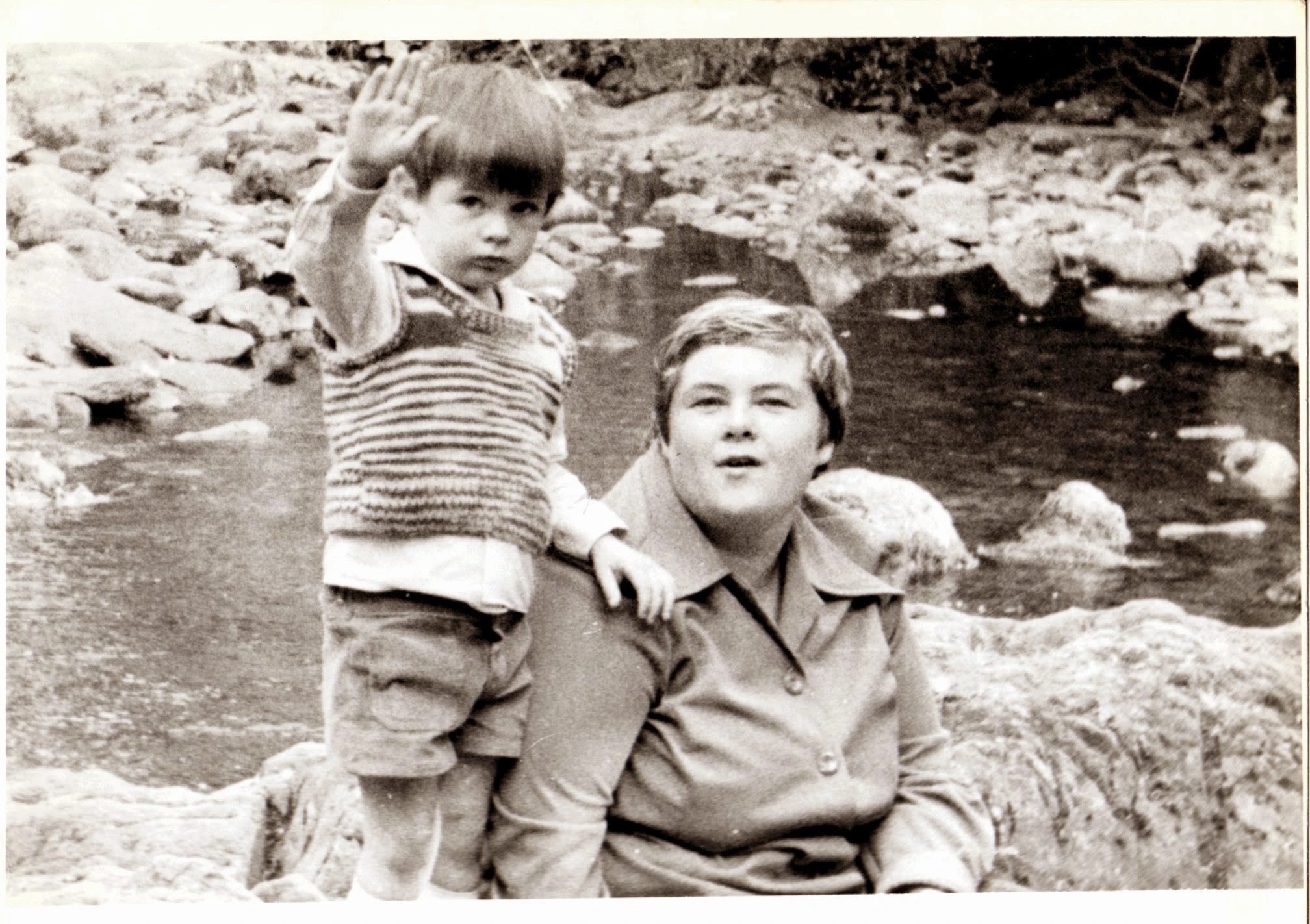 Black and white photo of Mum as a young woman by a stream, with a child (me)beside her waving at the camera.