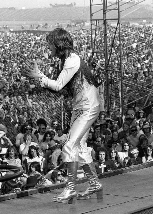 Ozzy Osbourne performing live at California Jam in 1974, wearing silver platform boots and fringe, surrounded by a massive crowd – tribute to Ozzy Osbourne.